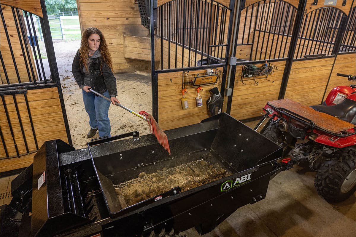 Woman mucking a stall in a barn to fill an ABI Attachments 25 Cubic Foot Ground Drive Manure Spreader