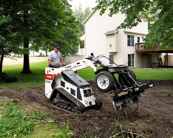 Mini SR being operated by a landscaper, with a large house and tall trees in the background.