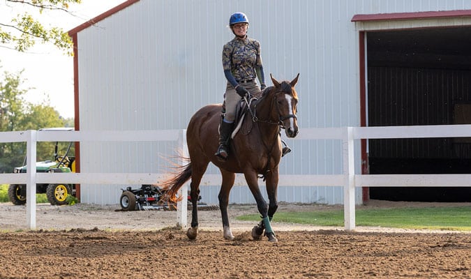 Female is riding a horse. The fencing is white and so is the barn in the background.
