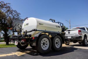 Side rear view of the ABI Attachments 550 DOT PRO Water Trailer as it is being towed by a pickup truck that is leaving a parking lot